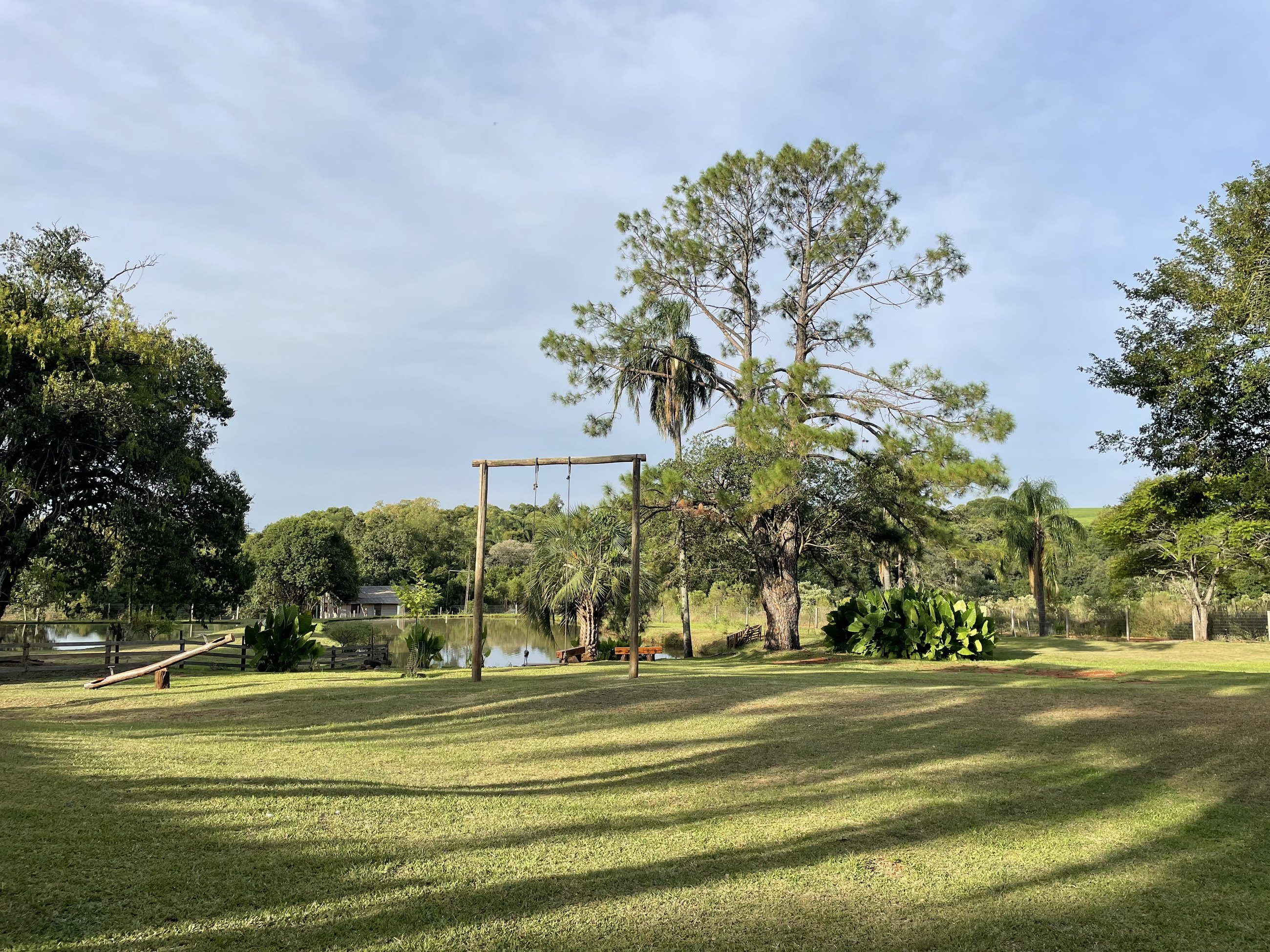 Gramado amplo com balanço e gangorra de madeira, árvores altas e o lago ao fundo
