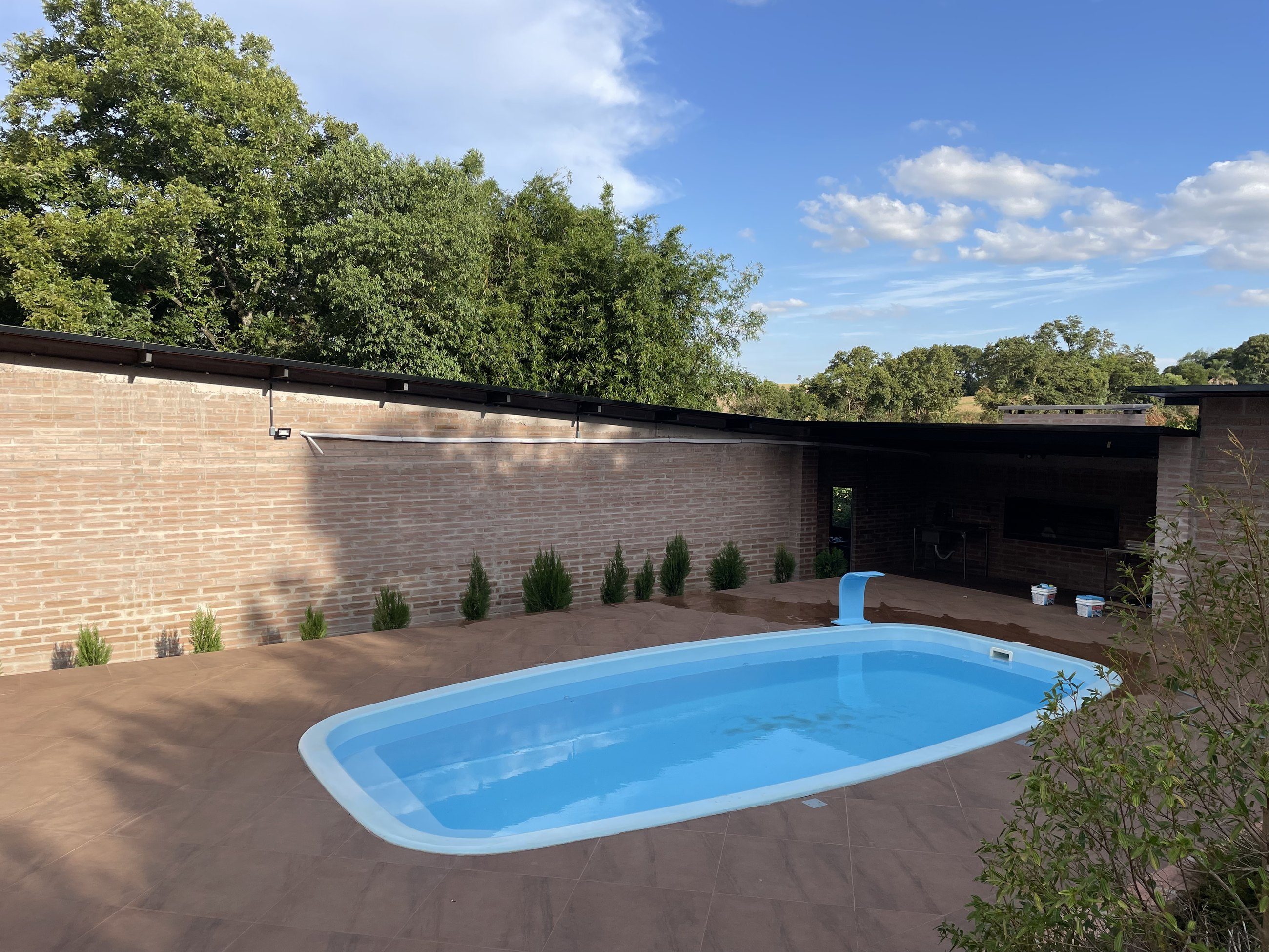 Piscina azul em deque cerâmico junto a um muro de tijolos aparentes, cercada por árvores