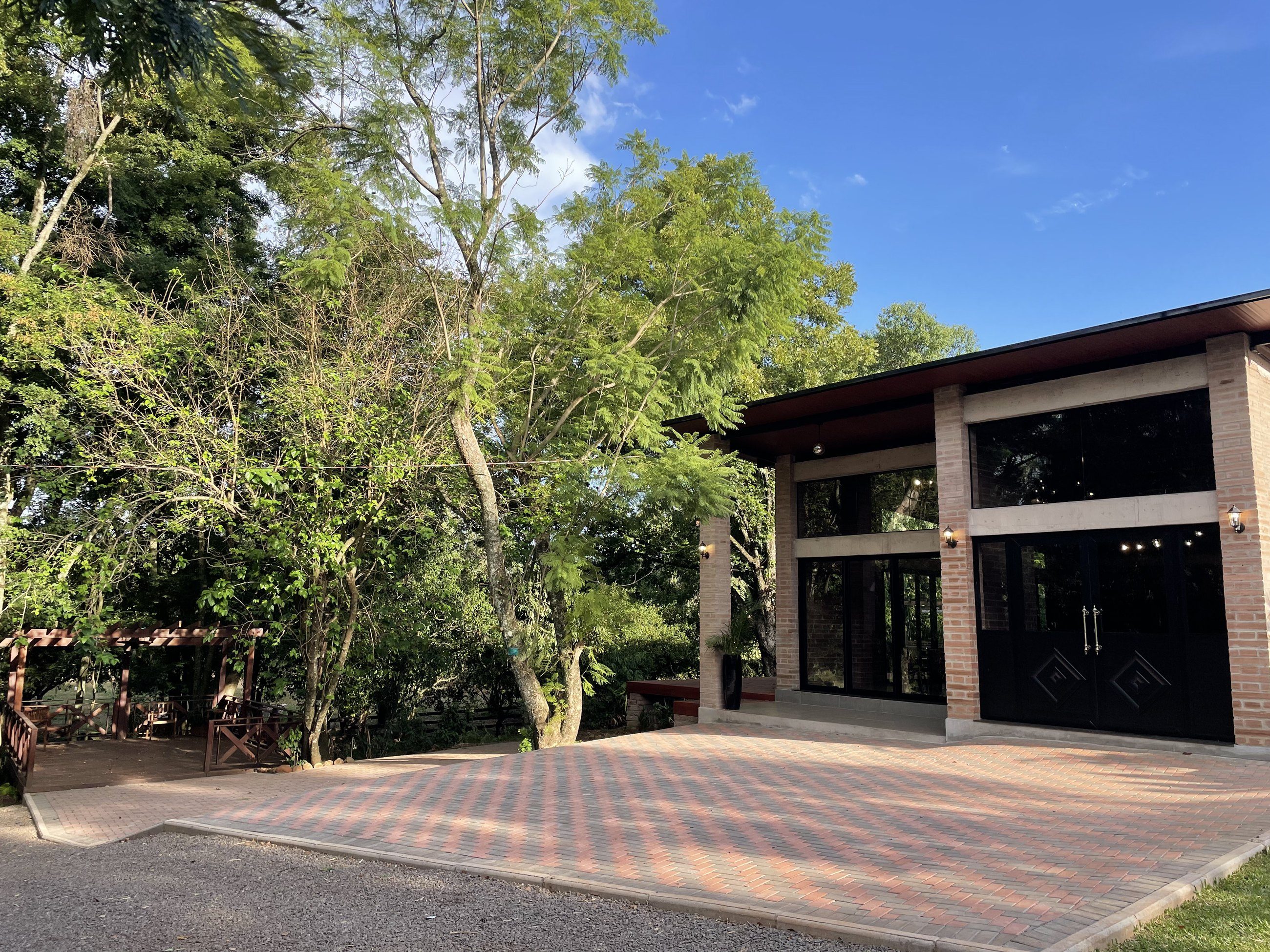 Fachada do salão de eventos com grandes portas de vidro e pátio de piso intertravado entre árvores